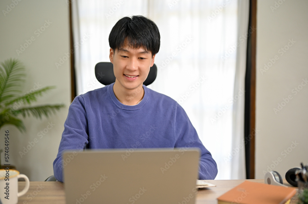 Handsome young Asian male college student in cozy sweater using laptop, at home. Handsome young Asian male college student in cozy sweater using laptop, at home.