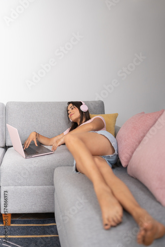 Young lady working on her laptop while listening to music on her headphones, lying on the couch