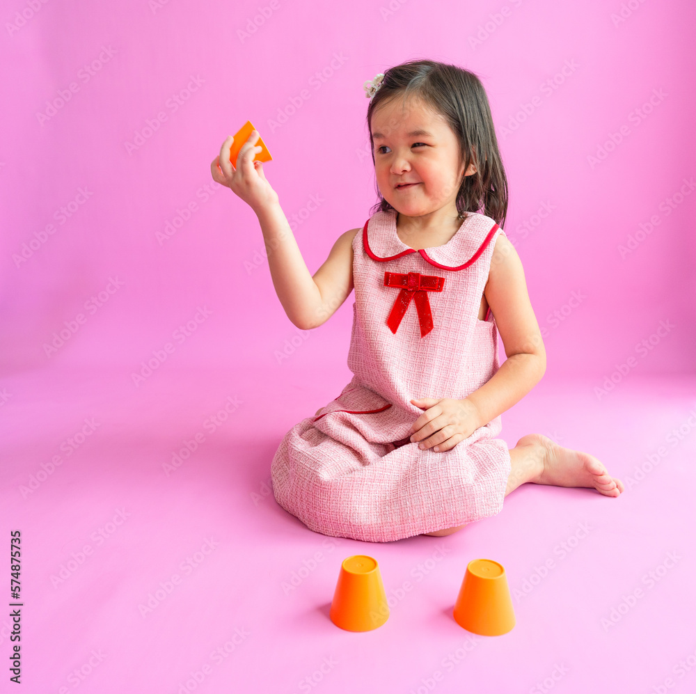 Pretty little girls sitting on floor and playing with three orange cups. She is trying to find the prize under one cup. Pretty little girls sitting on floor and playing with three orange cups. She is trying to find the prize under one cup.