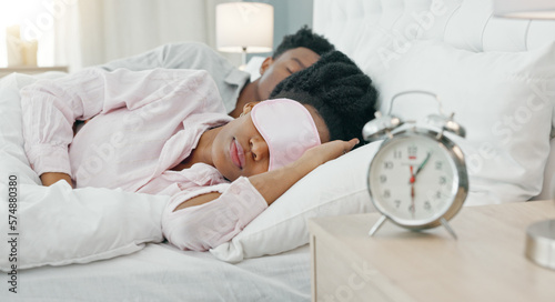 Black woman waking up in bed to switch off her alarm clock with a sleep mask in her house. Lazy, sleepy and African girl sleeping in her comfortable bedroom with an early morning get up time at home.