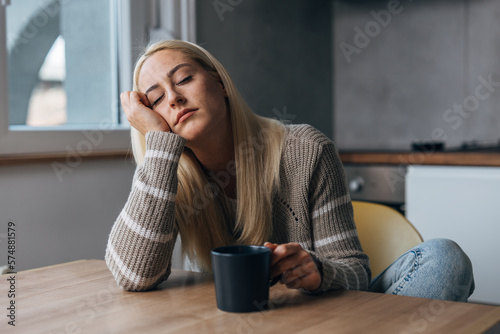 Tired Caucasian woman is sitting in the kitchen with a cup in her hand