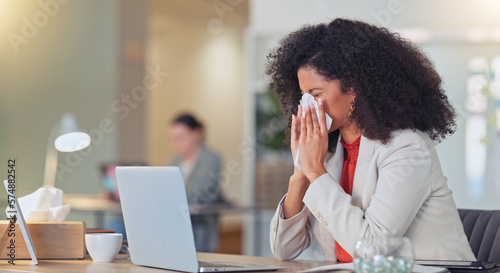 Sick businesswoman blowing nose with a tissue and suffering from flu virus or sinuses while working on a laptop in a modern office. Professional female with afro feeling ill while busy typing email
