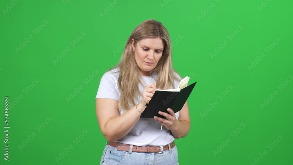 Focused woman writes down successful ideas in a notebook, an emotional blonde woman standing on a green background, plus size model, a chroma key template.