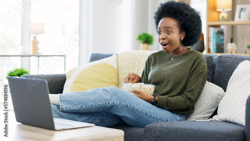 Fotografiet Happy african american woman streaming online movies on a laptop while snacking on popcorn and relaxing on a sofa at home