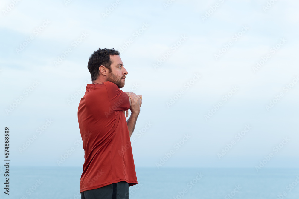 Side view of a male athlete doing stretching exercises in front of the sea.