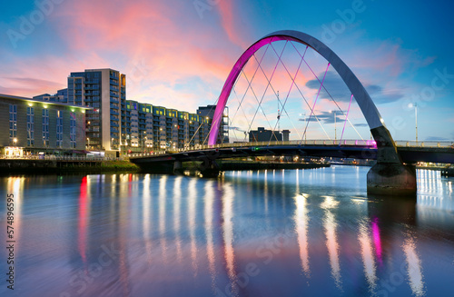 Fototapeta Naklejka Na Ścianę i Meble -  Beautiful Sunset Clyde Arc Bridge across river in Glasgow, Scotland, UK. It is nice weather with reflection on water, blue sky, lights from buildings in downtown, skyline, attractions.