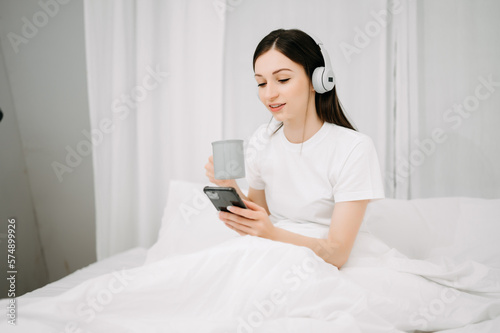 Beautiful young woman on a white bed with coffee cub and using smart phone at home. lifestyle concept.