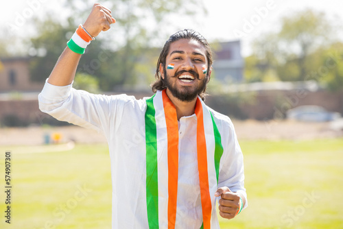 Portrait of cheerful young man fan wearing traditional white kurta and tricolor duppata with face painted cheering for indian sports team, celebrating Independence day or Republic day. photo