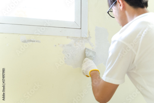 Asian painter man using scraper or spatula to remove old paint wall for cleaning, repair, preparing concrete wall before painting wall at home. Scraping to remove paint plaster peel. Painting concept.