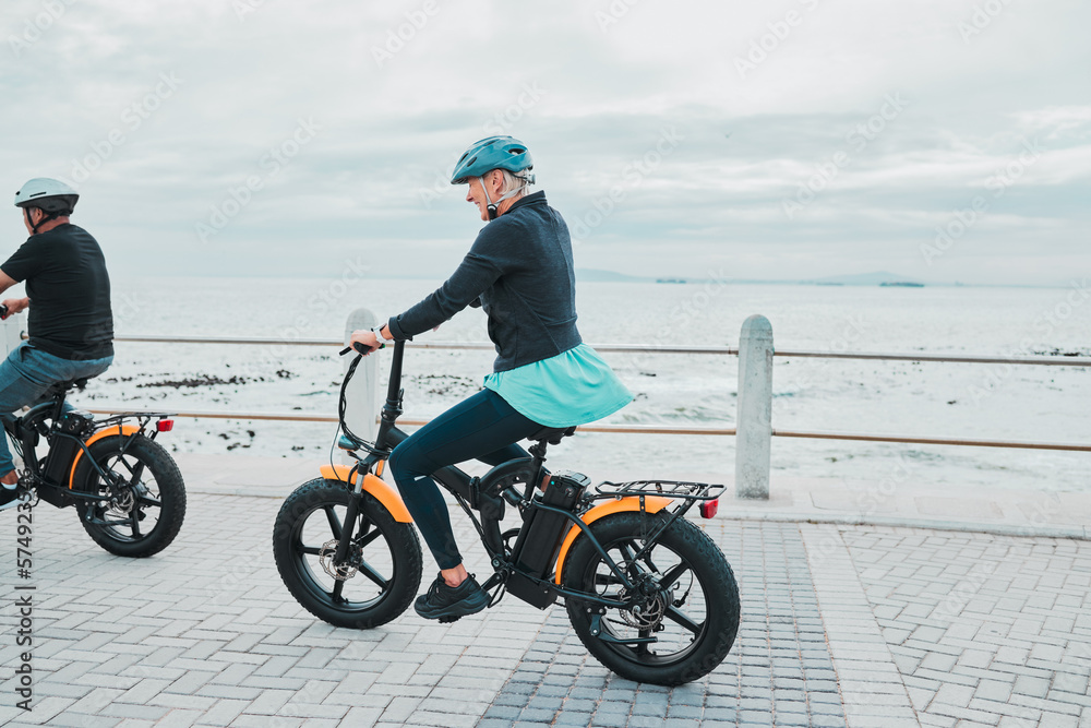 Fototapeta premium Couple, helmet or electrical bike by beach, ocean or sea in future transport, clean energy or sustainability travel. Technology, electric or eco friendly bicycle for happy woman or mature cycling man