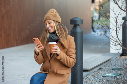 Photography Blonde woman chatting, browsing internet online on smartphone during coffee break while relaxing on park bench