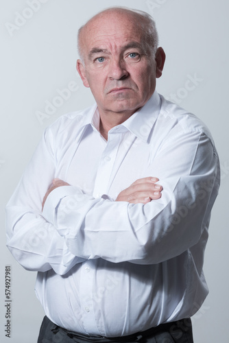 Confident senior man in white shirt crossing hands on chest and looking at camera while standing against gray background. Self confident senior isolated white studio shoot. © .shock