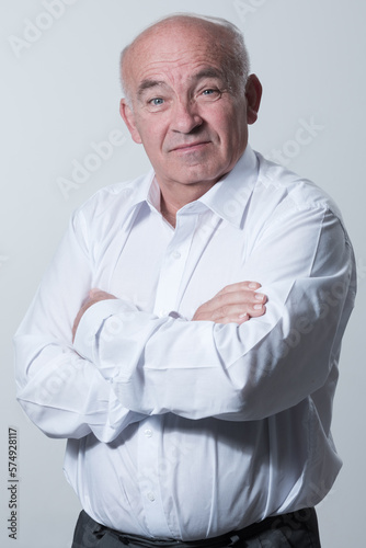 Confident senior man in white shirt crossing hands on chest and looking at camera while standing against gray background. Self confident senior isolated white studio shoot. © .shock