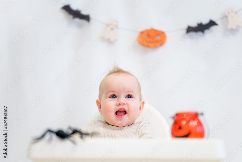 The baby toddler is sitting behind a high chair surrounded by Halloween attributes: pumpkins, spiders and garlands. Halloween Concept The baby toddler is sitting behind a high chair surrounded by Halloween attributes: pumpkins, spiders and garlands. Halloween Concept