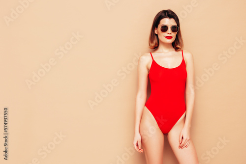 Young beautiful sexy woman. Smiling carefree model wearing red lingerie. Hot tanned female posing near beige wall in studio in summer swimwear bathing suit. In sunglasses.  slim and fit