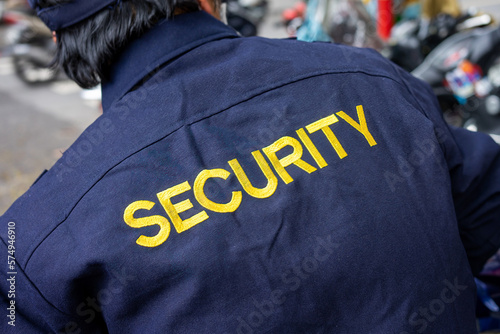 A security guard from behind with a blue jacket and the word security in yellow. Close up on a security guard's jacket.