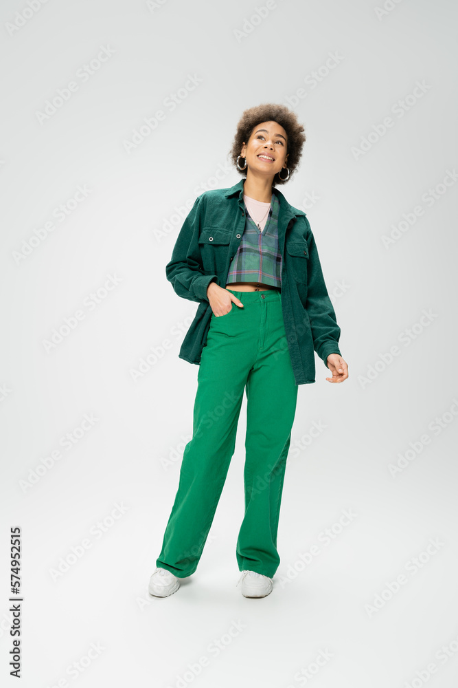 full length of joyful african american woman standing with hand in pocket of green pants and looking away on grey background. full length of joyful african american woman standing with hand in pocket of green pants and looking away on grey background.