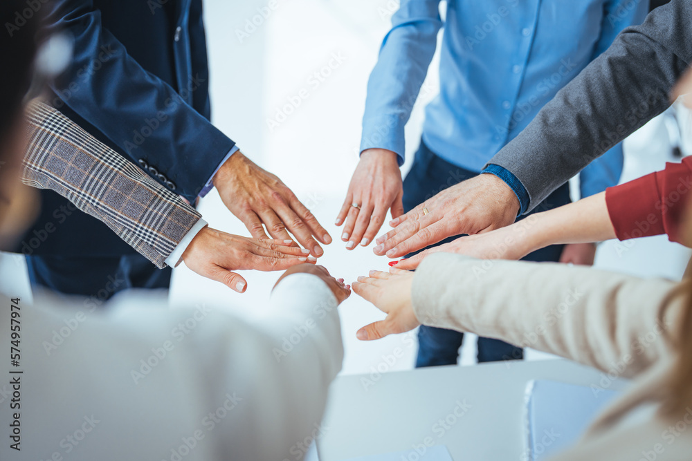 A high-angle shot of a group of male and female colleagues putting their hands together in an office. They are dressed in fashionable business clothes. Their faces are not visible, only their arms. 
