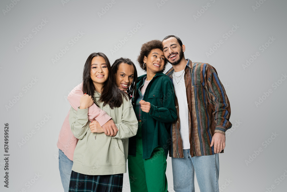 Fototapeta premium cheerful multicultural friends in stylish casual attire embracing while posing isolated on grey.