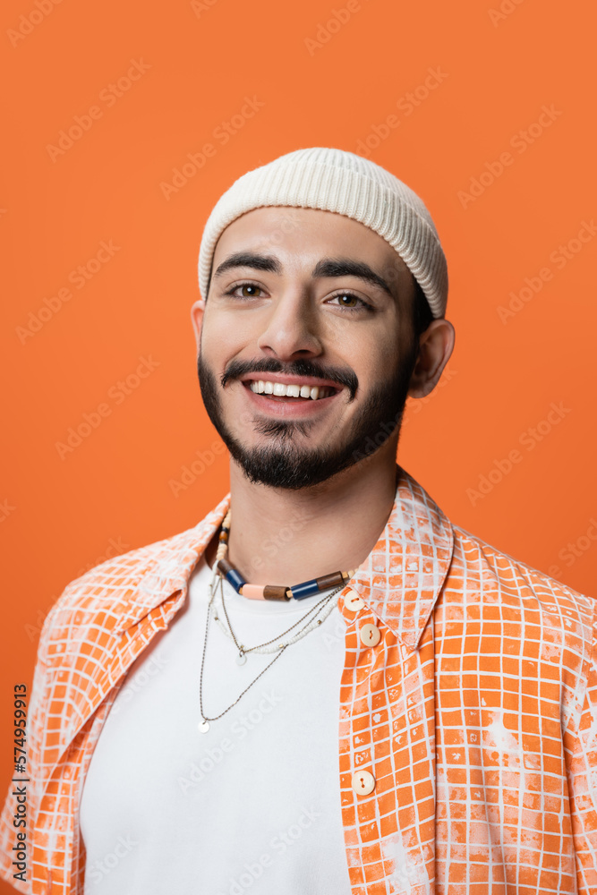 portrait of stylish bearded man in beanie and beads smiling at camera isolated on orange. portrait of stylish bearded man in beanie and beads smiling at camera isolated on orange.