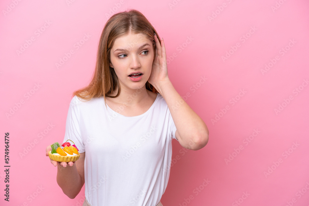 Teenager Russian girl holding a tartlet isolated on pink background listening to something by putting hand on the ear Teenager Russian girl holding a tartlet isolated on pink background listening to something by putting hand on the ear