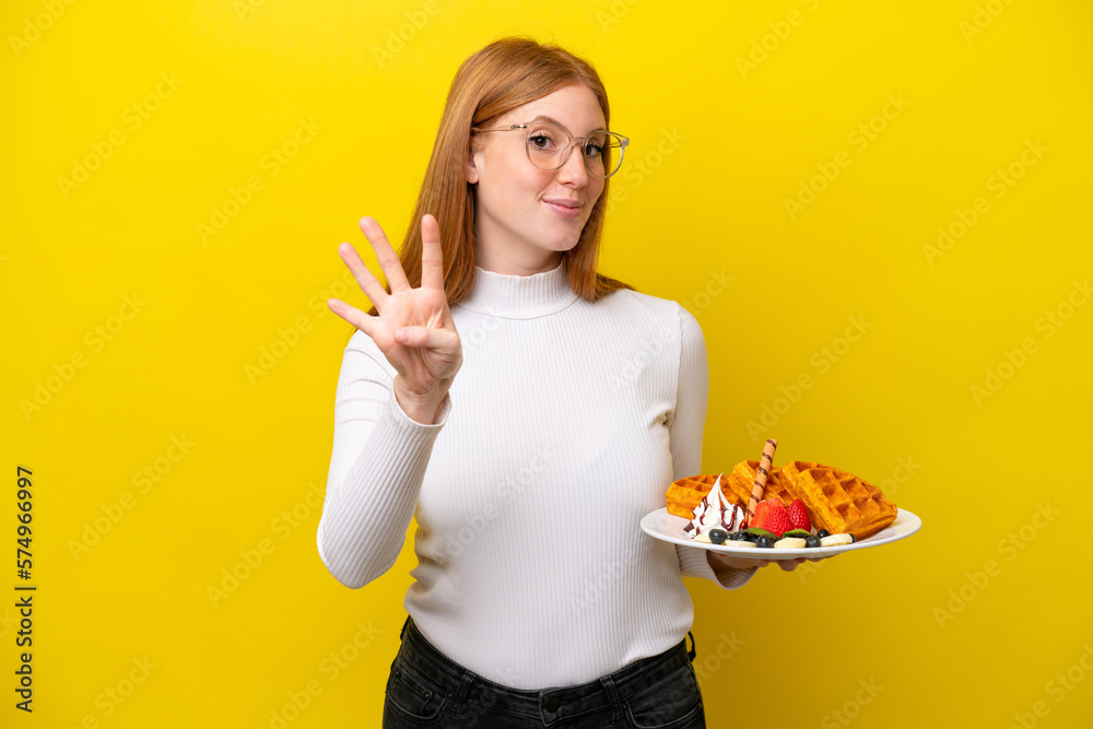 Young redhead woman holding waffles isolated on yellow background happy and counting four with fingers Young redhead woman holding waffles isolated on yellow background happy and counting four with fingers