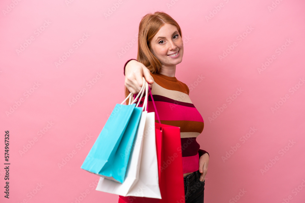 Young redhead woman isolated on pink background holding shopping bags and giving them to someone