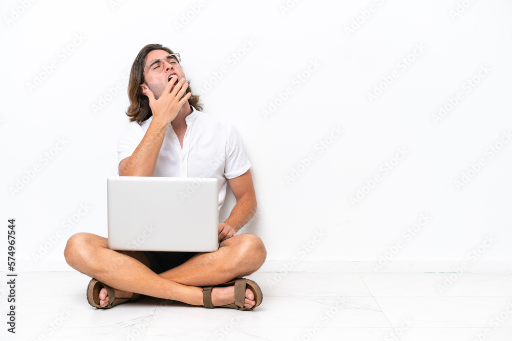 Young handsome man with a laptop sitting on the floor isolated on white background yawning and covering wide open mouth with hand Young handsome man with a laptop sitting on the floor isolated on white background yawning and covering wide open mouth with hand