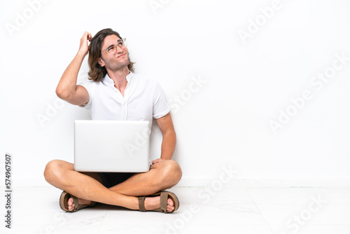 Young handsome man with a laptop sitting on the floor isolated on white background having doubts and with confuse face expression