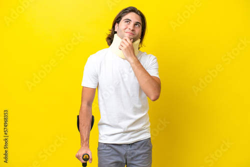 Young handsome man wearing neck brace and crutches isolated on yellow background and looking up