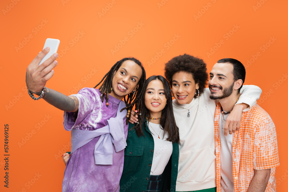 trendy man with dreadlocks taking selfie with happy multicultural friends isolated on orange. trendy man with dreadlocks taking selfie with happy multicultural friends isolated on orange.