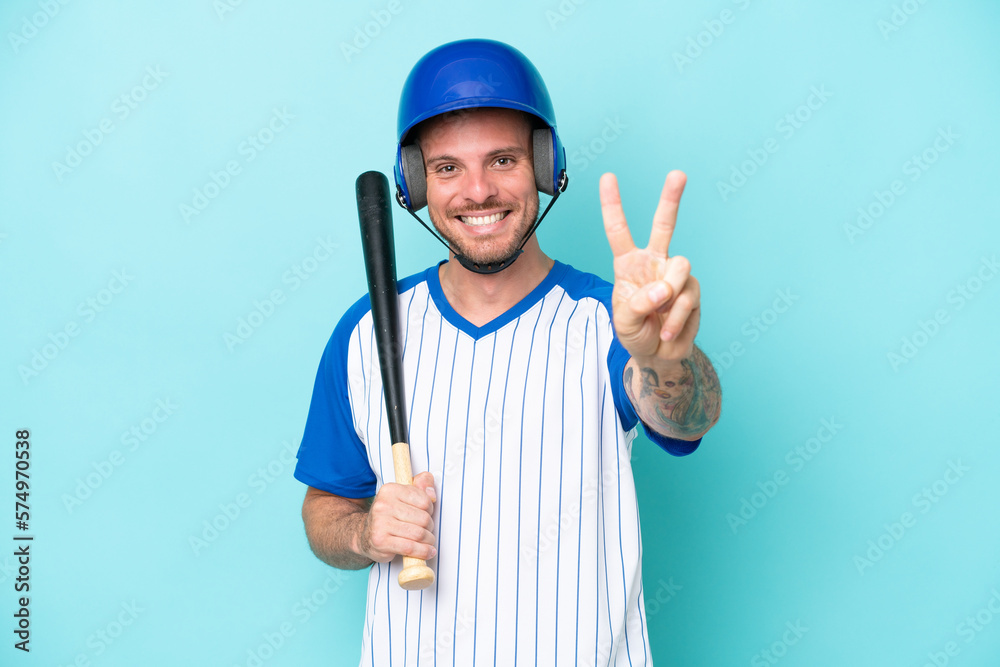 Baseball player with helmet and bat isolated on blue background smiling and showing victory sign Baseball player with helmet and bat isolated on blue background smiling and showing victory sign
