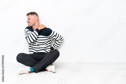 Young man sitting on the floor isolated on white background suffering from pain in shoulder for having made an effort