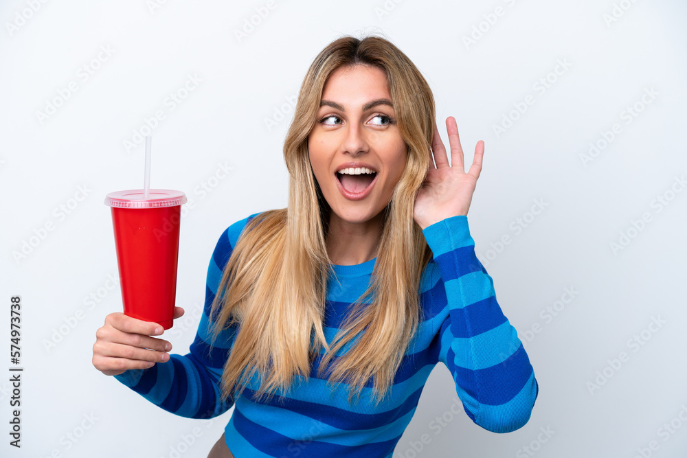 Young Uruguayan woman drinking soda isolated on white background listening to something by putting hand on the ear Young Uruguayan woman drinking soda isolated on white background listening to something by putting hand on the ear