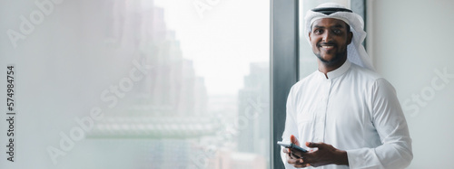Portrait of Successful Muslim Businessman in Traditional White Outfit Standing in His Modern Office, Using Smartphone Next to Window with Skyscrapers. Young Saudi, Emirati, Arab Businessman Concept.