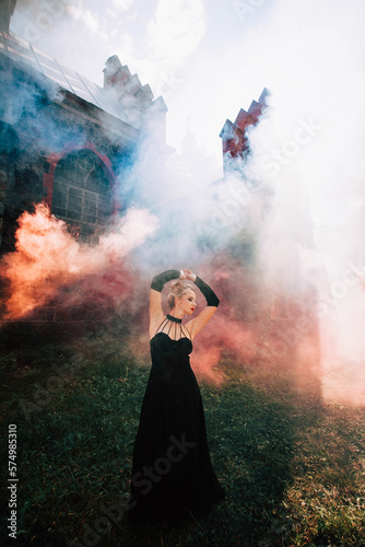 Art work. A slender beautiful woman with blond hair in a black vintage dress and expensive jewelry, stylish makeup and hairstyle, comes out of an ancient castle in fire and smoke.