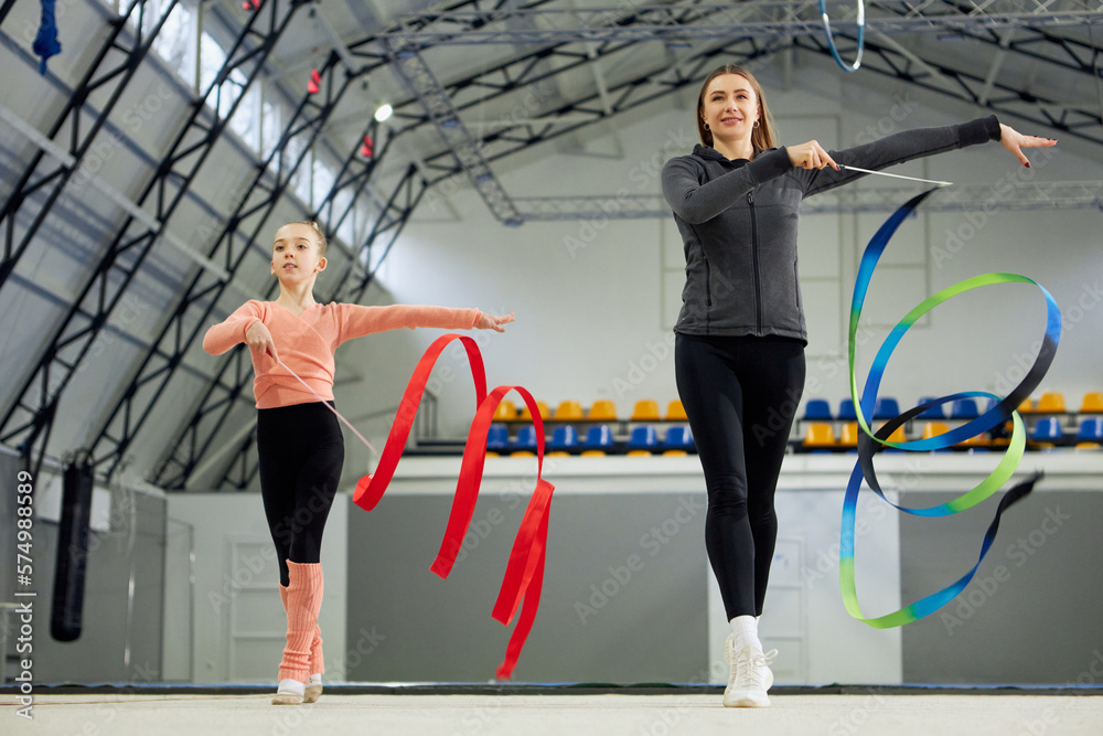 Fototapeta premium Little girl, child, rhythmic gymnast training indoors with coach, doing exercise with ribbon. Concept of sportive lifestyle, childhood, education, health, professional sport, championship