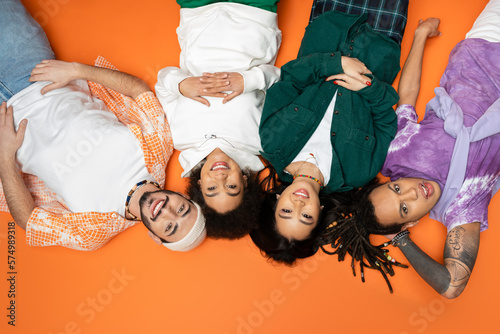 top view of joyful and stylish multicultural friends lying on orange background.