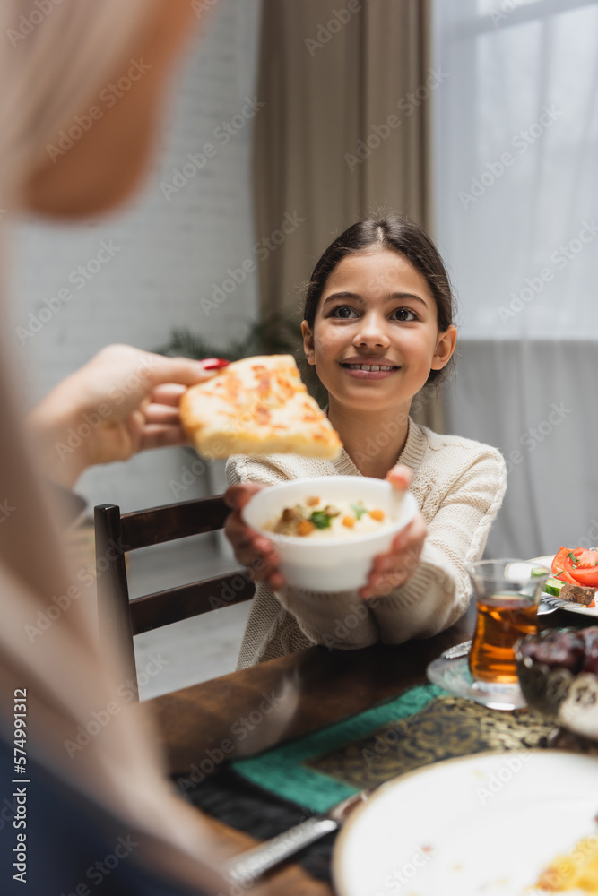 Positive muslim girl holding sauce near blurred mom with pita bread at home. Positive muslim girl holding sauce near blurred mom with pita bread at home.