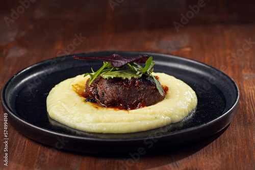 A medium rare ostrich steak in barbecue sauce on a cloud of herb mashed potato topped with fresh green arugula leaves on a black plate on a wooden background