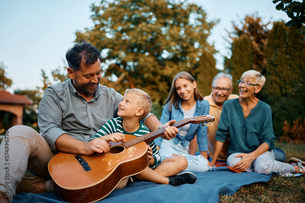 Happy father and son play acoustic guitar while relaxing with their family in backyard.