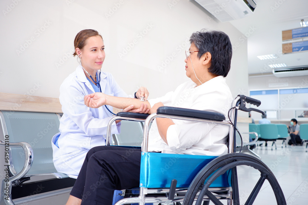 Female doctor using a stethoscope examining an elderly female patient sitting on a wheelchair inside a hospital medical concept.