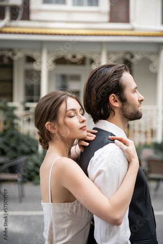 young bride in wedding dress standing with closed eyes and hugging groom in vest.