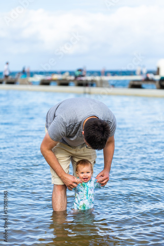 Happy baby standing in sea water in ocean pool at first trip to the beach with dad photo