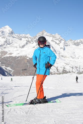 Skier in blue jacket, black helmet and orange pantson the piste slope in winter with snow mountains in Alps, Europe
