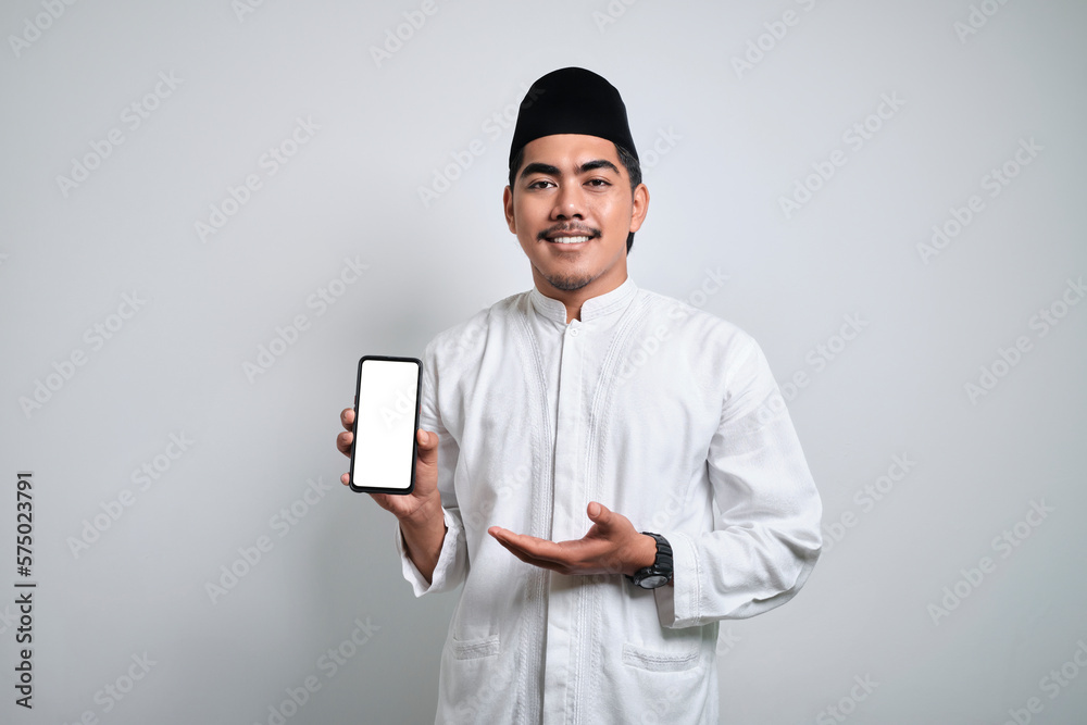Smiling young Asian Muslim man pointing finger at empty white display mobile phone screen isolated on white background Smiling young Asian Muslim man pointing finger at empty white display mobile phone screen isolated on white background