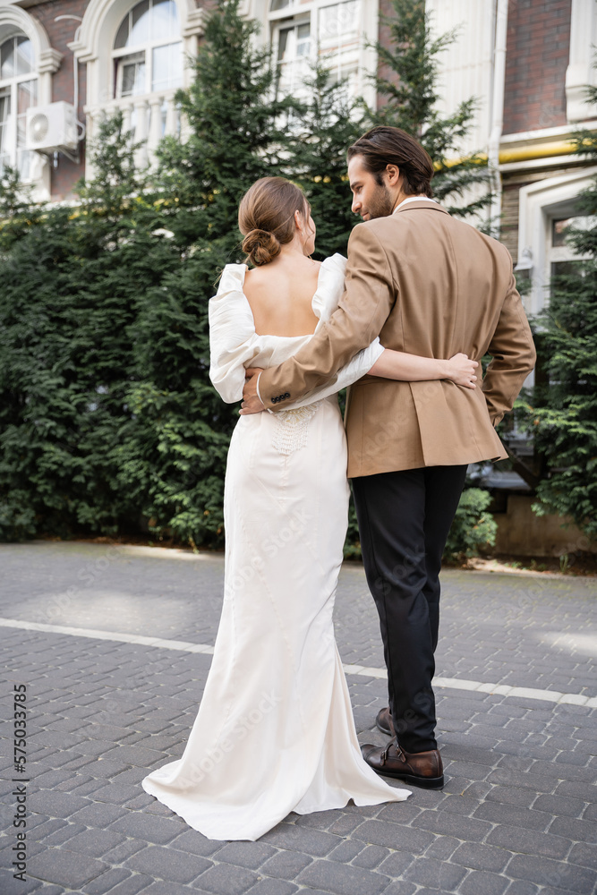 back view of bride in white dress hugging with bearded groom on street.
