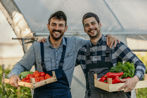 Portrait of two male workers standing with a crate full of fresh vegetables in front of a greenhouse. Small business concept. photo