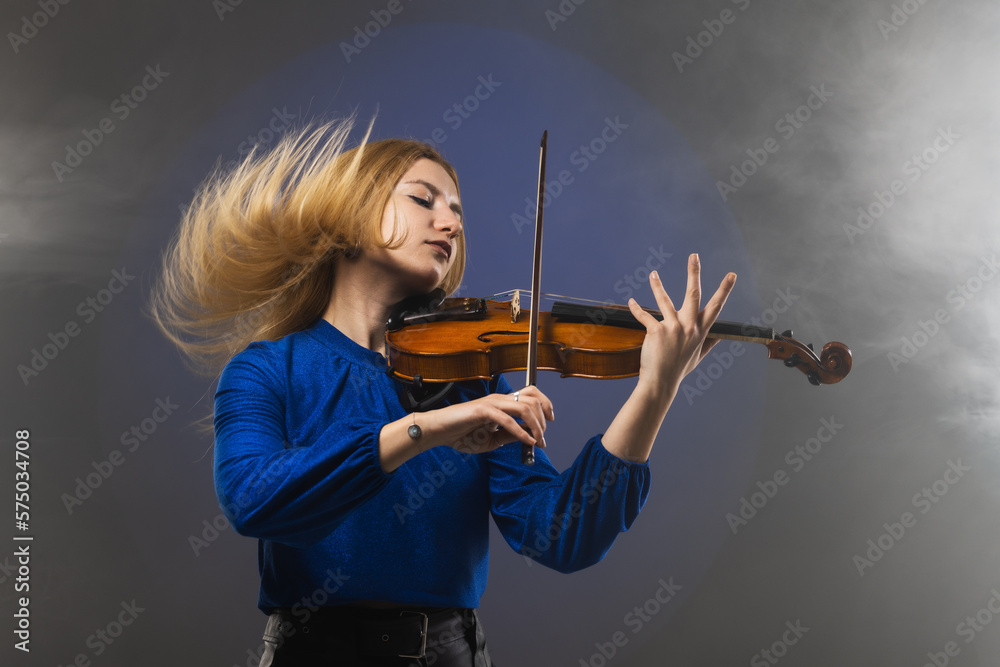 Beautiful blond girl playing violin. Female violinist doing energetic movement with hair in the air against blue background with smoke.