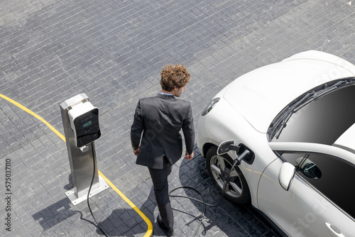 Fototapeta Aerial view of progressive businessman in black formal suit with his electric vehicle recharging battery at public car park charging station as vehicle powered by sustainable energy concept